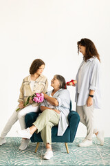 Cheerful european adult and teenager women give bouquet of flowers, gift to old grandmother
