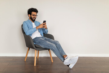 Mobile Communication. Indian Guy Messaging On Smartphone, Relaxing In Armchair At Home