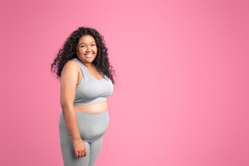 Portrait of body positive black lady in sports top bra and leggings posing at camera on pink...