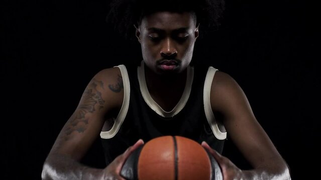 Basketball player sitting on a bench holding a ball. Concentrated african american sports man against black background