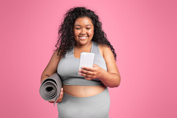 Smiling black chubby woman in sportswear with mat chatting on smartphone, standing on pink studio background