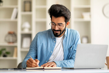 Smiling Indian Man Taking Notes While Working With Laptop At Home Office