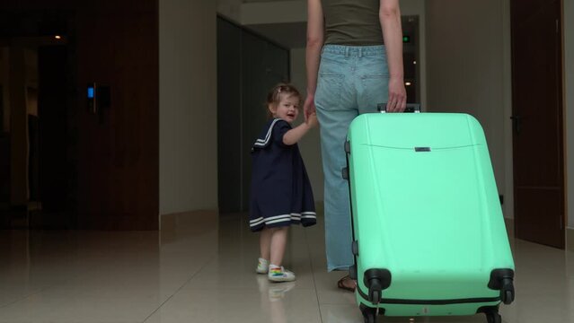A woman walks down a hotel corridor with her 3-year-old daughter, holding green travel luggage in her hand