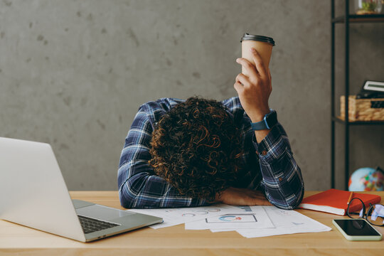 Tired young employee business Indian man he wearing casual blue checkered shirt hold takeaway delivery brown cup coffee to go put head on table sit work at office desk with laptop pc computer indoors.