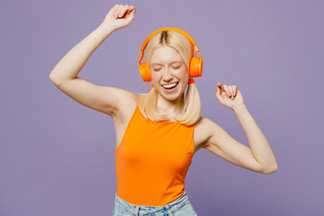 Young cheerful fun happy cool blonde woman wear orange tank shirt casual clothes listen to music in headphones raise up hands dance isolated on plain pastel light purple background. Lifestyle concept.