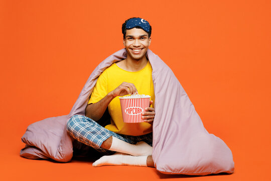 Full body young man in pyjamas jam sleep eye mask sit wrapped in duvet rest relax at home eat popcorn watch movie film isolated on plain orange background studio portrait Good mood night nap concept
