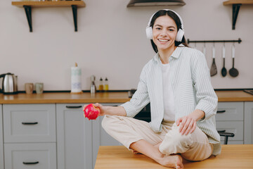 Young smiling happy housewife latin woman wear casual clothes striped shirt headphones listen to music eat apple look camera sit on table in light kitchen at home alone Lifestyle cooking food concept