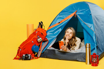 Full body young woman sit near bag with stuff tent using mobile cell phone isolated on plain yellow background. Tourist leads active lifestyle walk on spare time. Hiking trek rest travel trip concept.