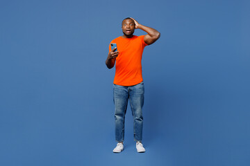 Fototapeta na wymiar Full body fearful young man of African American ethnicity he wear orange t-shirt hold head use mobile cell phone isolated on plain dark royal navy blue background studio portrait. Lifestyle concept.