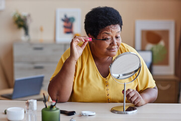 Mature woman in yellow pullover applying mascara on her eyelashes and looking in mirror while...