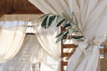 Closeup of green olive tree branch, blurred organza veils. White beach canopies, chuppah. Luxury beach tents at resort, gazebo. Mediterranean outdoor wedding concept. Summer birthday party, vacation.