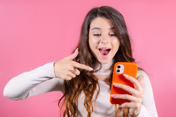 A woman brimming with excitement as she enthusiastically points her finger at a mobile phone, showcasing her enthusiasm for an exciting new app on the pink background 
