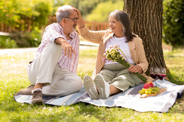 Cheerful senior european man gives bouquet of flowers to woman, enjoy picnic together in park, outdoor