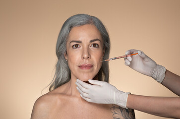 Attractive senior grey-haired woman getting cosmetic injection in nasolabial folds, standing on beige studio background