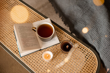 Cup of black tea stay on open paper book with scented candles on rattan table in living room top view. Selective focus. Good morning. - obrazy, fototapety, plakaty