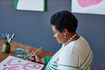Naklejka premium Mature female artist in casualwear painting with gouache or water colors on sheet of paper while sitting by desk in studio of arts and design