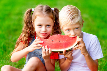 children girl and boy blonde in summer on the lawn with eating watermelon on the green grass having fun and rejoicing biting it, space for text