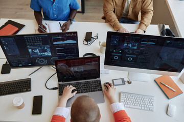 Above view of three intercultural coworkers analyzing cyber data on screens of their computers...