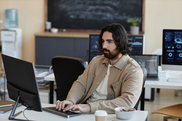 Young serious businessman typing on keyboard and looking at computer screen while analyzing data or...