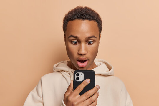 Horizontal shot of dark skinned man with curly hair has startled expression mouth wide open reflects his disbelief astonishment holds mobile phone dressed in sweatshirt isolated on beige background