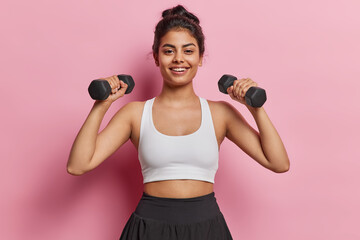 Horizontal shot of cheerful sporty slim woman doing exercises with dumbbells strengthening her body at home staying healthy smiles gladfully dressed in white top isolated over pink background - obrazy, fototapety, plakaty