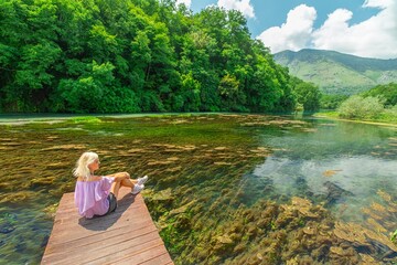 woman tourist enjoys the breathtaking beauty of the Blue Eye by sitting on a wooden platform built above the pool. Covered in lush greenery, trees and plants, creating a serene and picturesque setting