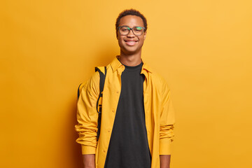 Portrait of delighted cheerful guy with short dark hair dressed in casual clothing poses with backpack has travel lifestyle poses against bright yellow wall feels slef proud after success on session