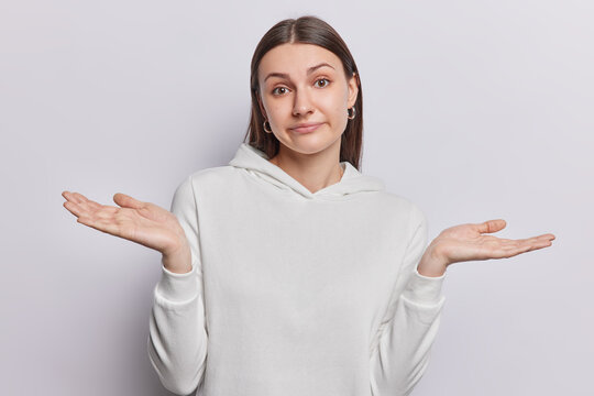 Clueless young dark haired woman has face filled with uncertainty spreads palms sideways cannot decide what to do wears casual sweatshirt isolated over white background hesitantly reaching decision