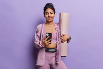 Young happy sporty girl holds mobile phone and rolled karemat smiles gladfully going to have pilates classes with instructor downloads app for sport poses indoor against purple stuio background.