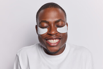 Headshot of dark skinned adult man applies white beauty patches under eyes to reduce wrinkles smiles toothily has eyes closed dressed casually isolated over white background. Beauty procedures