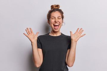 Waist up shot of positive cheerful woman raises her palms and exclaims loudly responding to...