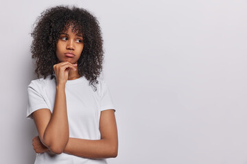 Fototapeta na wymiar Unhappy frustrated young African woman with dark curly hair keeps hand under chin looks sadly aside ressed in casual t shirt has bad mood dressed in casual t shirt isolated over white background