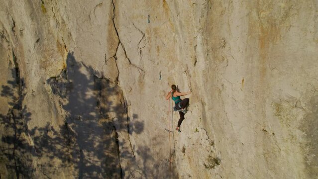 AERIAL: Female climber in the middle of a climb on a difficult climbing route. She is clipping in belay rope while lead climbing in limestone wall at incredible climbing playground in Karst landscape.