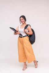 A whole body image of a smiling young Asian woman with backpack slung on one shoulder and holding a laptop another arm . Isolated on a white background.