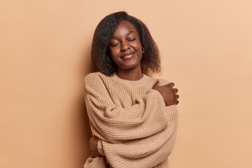 Joyful black woman in brown knitted sweater embracing herself with closed eyes has pleasant dreams...