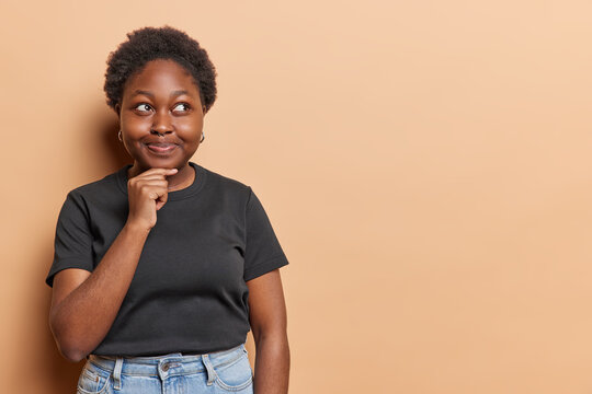 Thoughtful contemplative chubby young African woman keeps hand on chin focused aside has intention to do something dressed in black t shirt and jeans isolated over brown background. Let me think