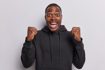 Photo of cheerful dark skinned woman clenches fists cheers over something exclaims loudly...