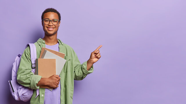 Clever male student prepares for exams points his index finger at blank space suggesting recommendation for academic education holds notepads poses with rucksack isolated over purple background