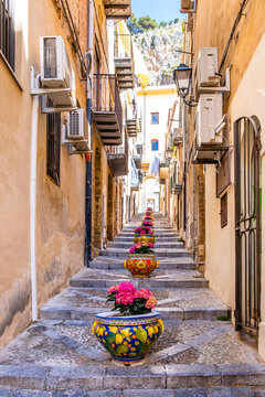 Narrow street in the old town of Cefalu, medieval village on Sicily island, Italy. Flower pots with traditional sicilian decoration. La Rocca cliff. Popular tourist attraction in Province of Palermo