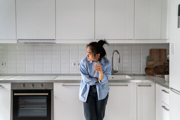 Unhealthy Asian woman suffering from strong sudden abdominal throbbing pain, standing in kitchen at...