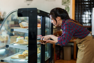 Asian female worker wearing apron holding bakery tongs wooden tray in hand, walk over refrigerated cake cabinet, open cabinet use cake tongs remove, places on wooden tray be served customers.