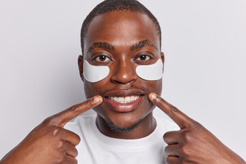 African dark skinned man takes care of his skin as he applies beauty patches under eyes to reduce bags and puffiness points at toothy smile showcasing results ofdedicated skincare routine poses indoor