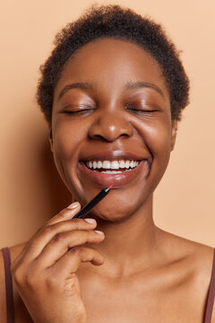 Vertical shot of dark skinned young woman applies lip pencil smiles broadly shows white teeth keeps eyes closed undergoes beauty procedures isolated over brown background doing makeup at home