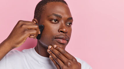 Sideways shot of serious dark skinned man meticulously massaging his face with gua sha stone has...