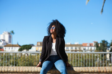 Young beautiful black woman with afro hair sitting on a bench wearing jeans and black jacket enjoying her holidays in spain. Travel and holiday concept.