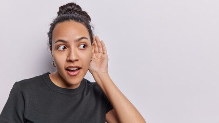 Fototapeta na wymiar Surprised curious Latin woman keeps hand near ear tries to overhear listening intently concentrated aside has widely opened eyes and mouth wears casual black t shirt isolated over white background
