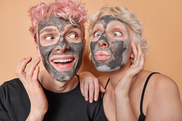 Horizontal shot of woman and man have fun apply beauty clay mask on faces for reducing pores and blackheads dressed in black t shirts smile happily isolated over brown background. Skin care procedures