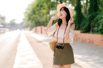 Portrait of asian young woman traveler with weaving hat and basket and a camera standing by the street. Journey trip lifestyle, world travel explorer or Asia summer tourism concept.