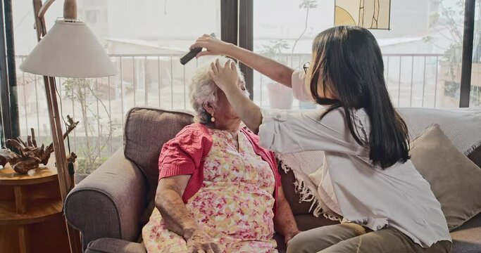 Cute latina teen brushes her lovely grandmother's curly hair with love and affection sitting on the couch in the living room while both spending a good time.