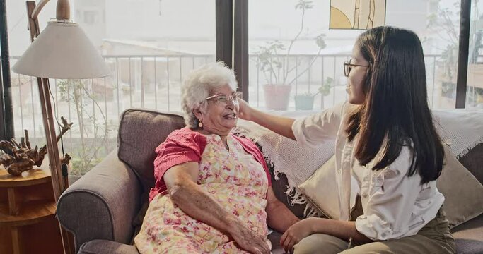 Cute Latina teen fixes her Latina grandmother's hair with care and love while spend a good time together sitting on the couch in the living room.